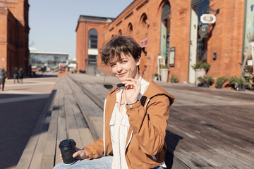 Young beautiful girl with haircut sitting on the street against a red brick wall and drinks coffee.