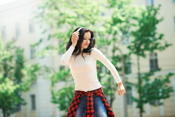 Street hip hop dancer on sunny day. Young happy sportive woman in urban background listening music in white wireless headphones and dancing. Hair fluttering in the wind