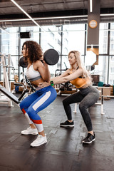 Fototapeta na wymiar Slender girl in stylish bright sports clothes doing back squats with barbell and another athletic girl helps her in the modern gym