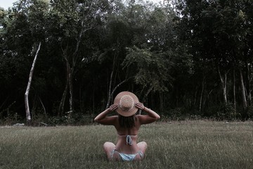 woman wearing bikini sitting on grass field holding hat facing forest