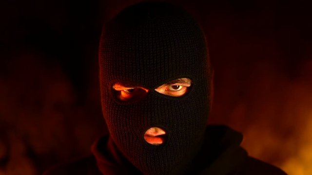 Portrait of young man in black balaclava against backdrop of a blazing night fire. Concept of mass rallies and riots