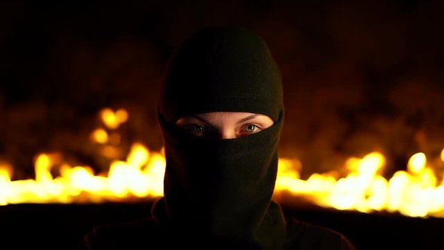 Portrait of female protesting activist in black balaclava against burning barricades at night. Concept of strikes, political conflicts and confrontation.