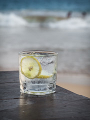 Glass of gin with tonic, lemon and ice cubes on the wooden table. Sea on the background.