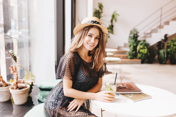 Graceful young lady resting at the table with glass of cold tea and magazines. Indoor portrait of...