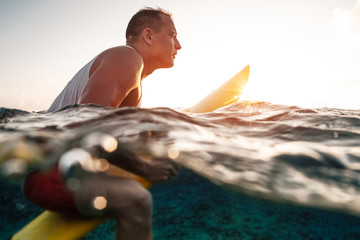 Splitted shot of the male caucasian surfer sitting on his surfboard in the ocean at sunset