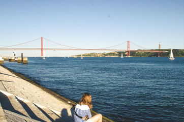 Woman sitting near river and looking to red 25 april bridge in Lisbon