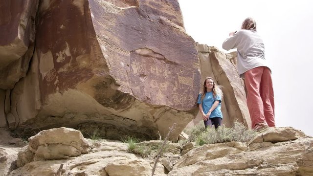 Woman standing next to petroglyphs as another takes picture as she stands next to the panel in Nine Mile Canyon in Utah.