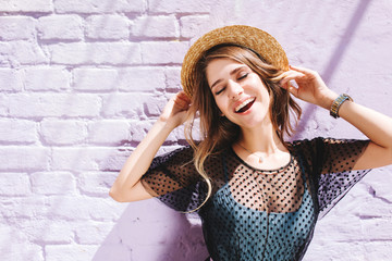 Amazing long-haired girl in stylish clothes enjoying good day outside standing with eyes closed under sun. Outdoor portrait of inspired lady in straw hat and wristwatch posing with happy smile.