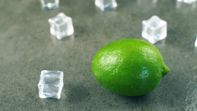 Macro shot of fresh green lime rotating on dark surface with crystal ice cubes. 4K close up footage.