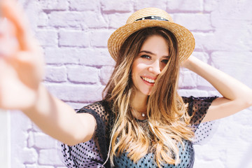 Close-up portrait of happy girl with mid-back length hair making selfie and holding straw hat. Outdoor photo of laughing romantic woman taking picture of herself in summer morning.