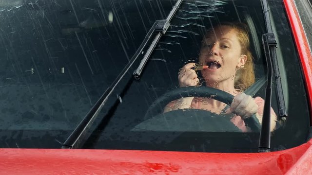 woman drives a car and does makeup at the same time. girl behind the wheel of a car paints her lips creating an emergency on the road. It is raining outside.