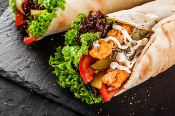 Tortilla wraps with grilled chicken fillet, fresh vegetables and salad on black stone background....