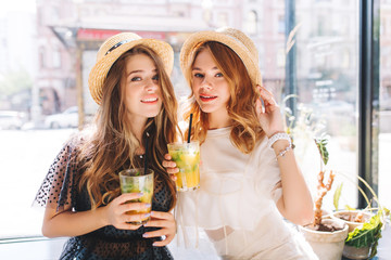 Close-up portrait of tired but happy girls enjoying cocktails after shopping in weekend. Stunning ladies in vintage attires relaxing together in cafe and drinking juice.