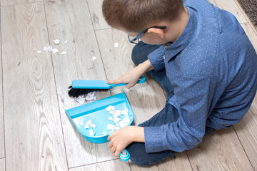 Chłopiec w okularach sprząta mieszkanie. Zamiata papierki na szufelkę za pomocą zmiotki. - obrazy, fototapety, plakaty