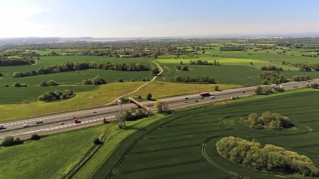 Forward moving aerial views across British countryside. M62 motorway traffic passing across both directions at speed. Blue sky, sunny farmland scenery.