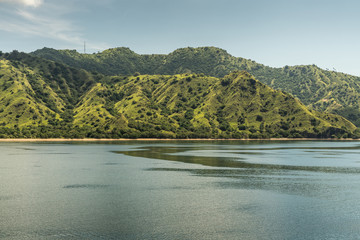Fototapeta premium Rinca Island, Indonesia - February 24, 2019: Closeup of hills and beaches on Westside coast in Savu Sea. Green hills, antenna tower in distance and flat sea.