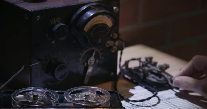 A cold war era spy sending messages via Morse Code using a code key with tape recorder going in the foreground.
