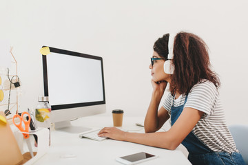 Slim black girl in headphones looking at computer's screen with attention. Indoor portrait of young...