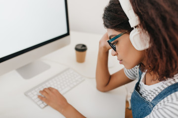 Close-up portrait of tired black girl in big white headphones sitting at workplace and typing on keyboard. Indoor photo of sad young woman with dark hair working with computer in her office.