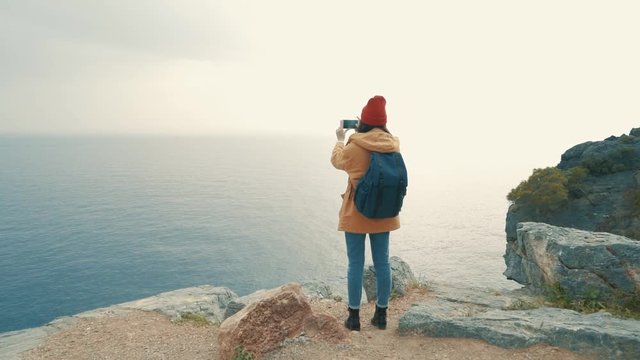 Tourist girl with a backpack standing on the edge of a cliff and shooting photo of the sea on an autumn cloudy day. Slow motion.