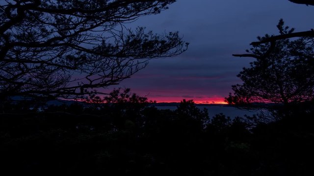 Time Lapse of sunset west coast of Norway. Trees surrounding the sides and foreground. Red dramatic color in the horizon.