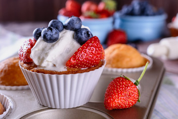 Homemade freshly baked muffins and one of them is decorated whipped cream and slices of strawberry and a few blueberries on top are in baking dish on a wooden table next to cream and berries 