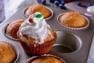 Homemade freshly baked muffins and one of them is decorated whipped cream on top are in baking dish on a wooden table next to cream and berries for decorating