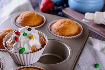 Homemade freshly baked muffins and one of them is decorated whipped cream on top are in baking dish on a wooden table next to cream and berries for decorating