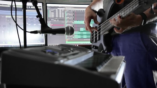 Musician playing bass guitar in a home studio with PC screens displaying software