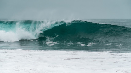 A large barreling wave crashing onto the shore spraying mist