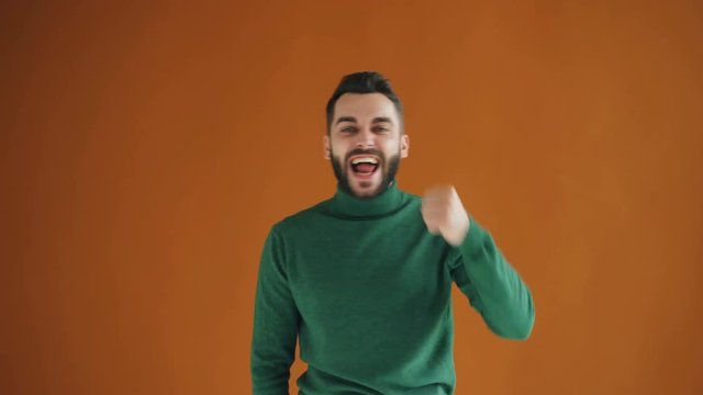 Portrait of excited young man raising fists celebrating success and luck expressing positive emotions standing against orange background. People and excitement concept.