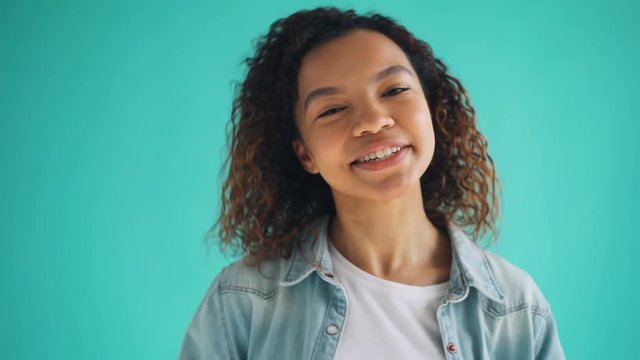 Portrait of cute African American teenage girl making funny faces and laughing enjoying silly activity standing on blue background. Youth and fun concept.