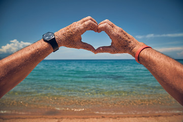 Man making heart shaped symbol with his hands on a sandy tropical beach.