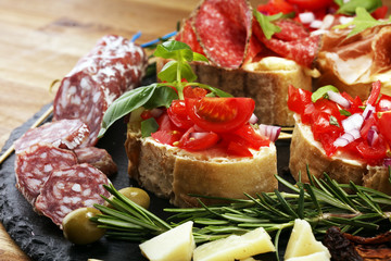 Traditional italian antipasto bruschetta appetizer with cherry tomatoes, cream cheese, basil leaves and balsamic vinegar on cutting board with prosciutto, salami, cheese