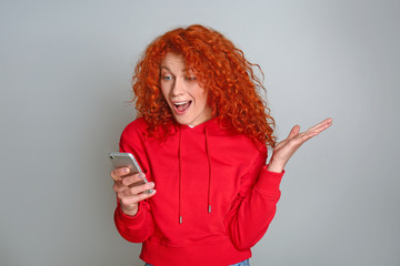 Surprised redhead woman with mobile phone on grey background