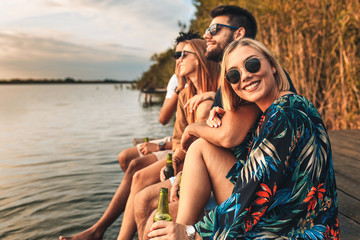 Group of friends enjoying a day at the lake. They sitting on pier talking, laughing and drinking beers. - obrazy, fototapety, plakaty