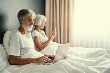 Happy family. Cozy home and technology. Beautiful senior woman and her husband using laptop computer and smartphone in bed.