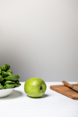 plate with spinach leaves near green apple and cutting board on grey