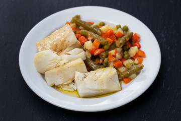 boiled cod fish with boiled vegetables on white plate on black ceramic background