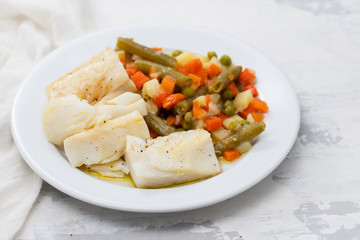 boiled cod fish with boiled vegetables on white plate on ceramic background