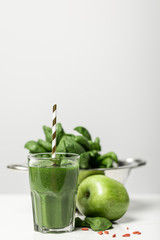 selective focus of green smoothie in glass with straw near fresh spinach leaves and apple on grey