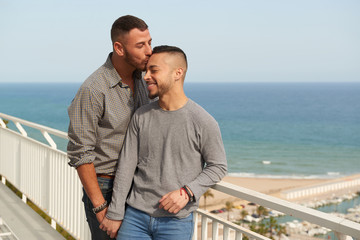 Una pareja gay en el balcón de su casa en un día soleado de verano con el puerto detrás.