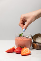 cropped view of woman adding coconut flakes in tasty smoothie near red strawberries on grey