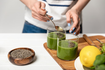 cropped view of woman holding spoon with chia seeds near glasses of green smoothie on grey