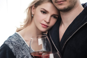 A couple, a man and a woman in bathrobes drink red wine from large glass goblets in the apartment. Kiss and hug.