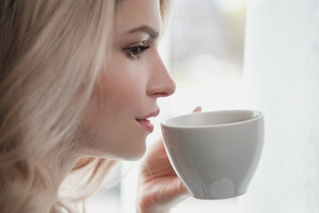 Beautiful young blonde woman in a blue robe by the window. Drinks coffee or tea from a white cup with a saucer. Morning, sunshine, bedroom window.