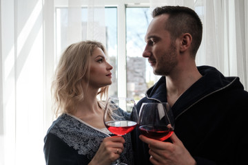 A couple, a man and a woman in bathrobes drink red wine from large glass goblets in the apartment. Kiss and hug.