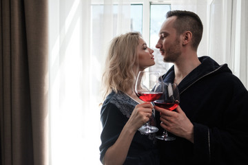 A couple, a man and a woman in bathrobes drink red wine from large glass goblets in the apartment. Kiss and hug.