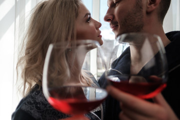 A couple, a man and a woman in bathrobes drink red wine from large glass goblets in the apartment. Kiss and hug.
