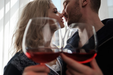 A couple, a man and a woman in bathrobes drink red wine from large glass goblets in the apartment. Kiss and hug.
